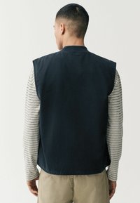 Marineblauw mouwloos vest met een gestructureerd ontwerp, gemaakt van katoen, gedragen over een beige en wit gestreepte langemouwen shirt. Minimalistische stijl.