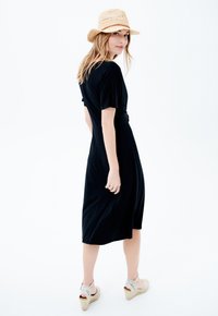 Robe midi noire à manches courtes et taille nouée, associée à des sandales espadrilles beiges et un chapeau en paille tressée. Texture simple et lisse.