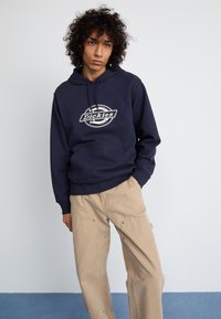 Mornarska dukserica s kružnim bijelim logotipom "Dickies" na prsima, u kombinaciji s bež cargo hlačama. Čini se da je materijal mekan i opuštenog kroja.