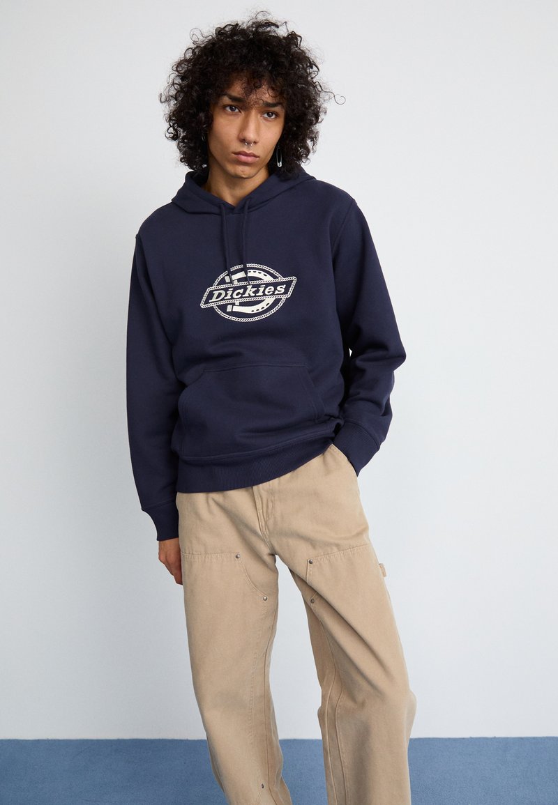 Felpa navy con un logo circolare bianco "Dickies" sul petto, abbinata a pantaloni cargo beige. Il tessuto appare morbido con una vestibilità comoda.