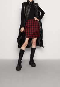 Gilet lungo in finta pelle nera, top nero, gonna mini a quadri rossi, borsa nera trapuntata e stivali alti fino al ginocchio neri con lacci.