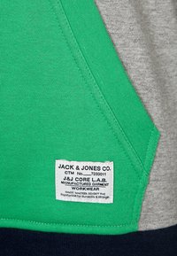 Tessuto verde e grigio con una finitura testurizzata. Presenta un'etichetta del marchio con la scritta "JACK & JONES CO." insieme alle istruzioni per la cura e ai dettagli sul lavoro.
