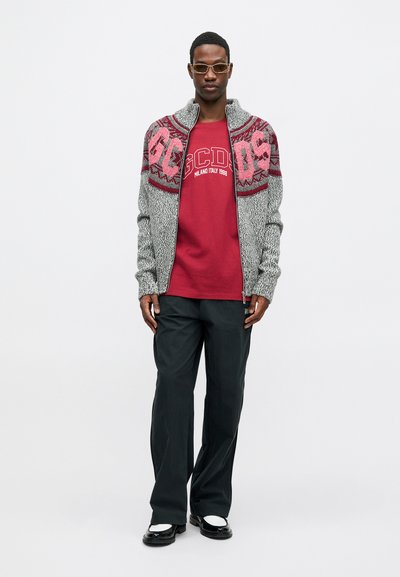 Grijs en rood gebreid vest met hoge kraag over een bordeaux logo t-shirt. Zwarte wijde broek en zwart-witte schoenen.