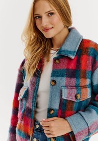 Pestrofarebný károvaný vlnený sakó s modrým golierom, hnedými gombíkmi a vreckami. Mäkká textúra s výraznými červenými, modrými a oranžovými časťami.