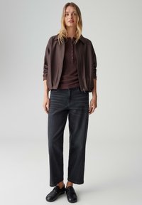 Braune Bomberjacke mit Reißverschluss über einem dunkelburgunder Henley-Shirt, kombiniert mit locker sitzenden dunklen Jeans und schwarzen Loafers.