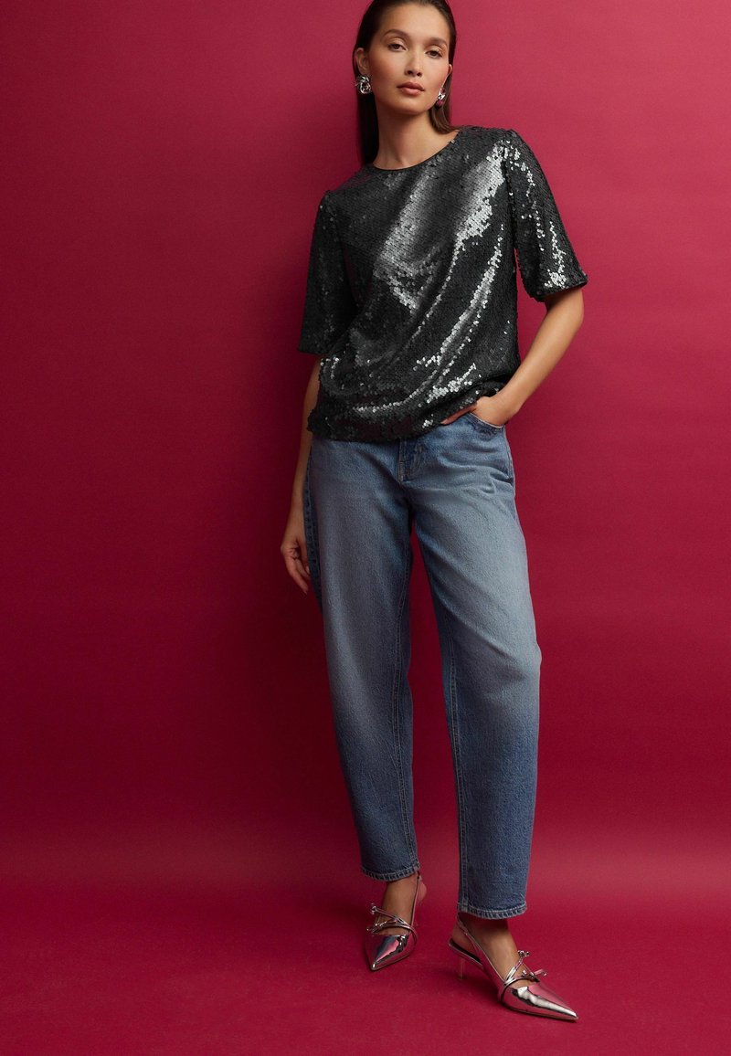 Haut en sequins noir à manches courtes, assorti à un jean ample bleu clair et des chaussures à talons argentées sur fond rose.