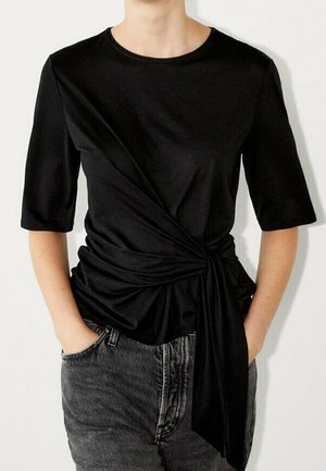 Henkilö, jolla on musta, lyhythihainen yläosa, jossa on vyötäröllä kerätty solmu, yhdistettynä haalistuneisiin mustiin farkkuihin.