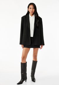 Sort blazer over en hvid polo, tekstureret sort nederdel og knæhøje sorte støvler med spidse tå. Modellen står imod en hvid baggrund.