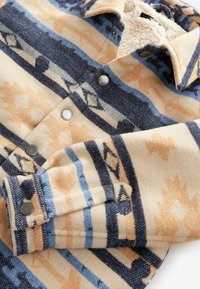 Wollen jas met beige, blauwe en zwarte geometrische patronen, drukknopen en een crème kleurige sherpa voering zichtbaar aan de binnenkant van de kraag.