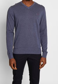 Pull à col en V à manches longues pour homme en tissu tricoté bleu foncé, avec poignets et ourlet côtelés, et un petit logo sur la poitrine.