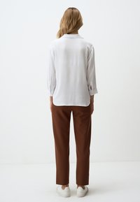 Blouse blanche à col avec une finition texturée, associée à un pantalon marron ajusté. Le bas de la blouse est légèrement arrondi ; les manches sont retroussées au niveau des poignets.