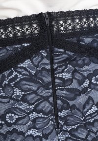 Svart spetsplagg med blommönster, en tunn dragkedja på mitten och en texturerad spetskant upptill. Mjukt tygbakgrund synlig.