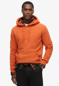 Sudadera con capucha naranja con un bolsillo frontal, combinada con una camisa de cuadros negra y naranja debajo, usada con pantalones oscuros y un reloj.