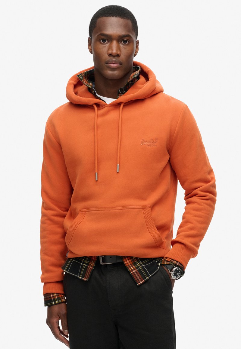 Sudadera con capucha naranja con un bolsillo frontal, combinada con una camisa de cuadros negra y naranja debajo, usada con pantalones oscuros y un reloj.