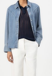 Personne portant une chemise en denim bleu par-dessus un polo en maille bleu marine, associée à un pantalon blanc à jambes larges, une main dans la poche.