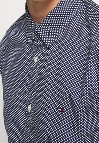 Camicia button-up blu navy scuro con un motivo geometrico bianco, dotata di un colletto classico e di una piccola etichetta con il marchio in rosso, bianco e blu.
