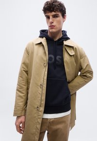 Νεαρός άνδρας που φοράει μπεζ καμπαρντίνα πάνω από σκούρο μπλε φούτερ με κουκούλα και ανοιχτό καφέ παντελόνι, στέκεται μπροστά σε απλό ανοιχτόχρωμο φόντο.