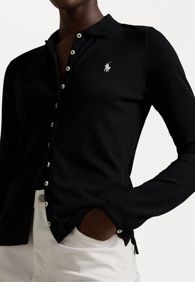 Black long-sleeve polo shirt with a collar, featuring white buttons and an embroidered logo on the left chest. Worn with white pants.