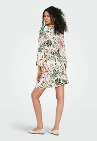 Blumenkleid mit weißem Hintergrund, versehen mit bunten Pflanzenmustern, Glockenärmeln und einer taillierten Taille. Kombiniert mit hellen Spitzenschuhen.