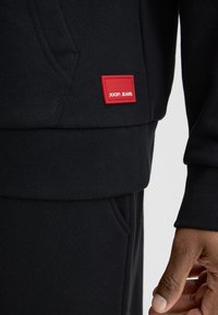 Felpa nera con tessuto testurizzato, tasca a marsupio e etichetta rossa "JOOP! JEANS" sul lato sinistro inferiore.