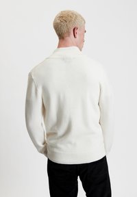 Maglione bianco lavorato a maglia con colletto e polsini a coste, caratterizzato da una superficie liscia e una vestibilità comoda. Modello visto da dietro, abbinato a pantaloni neri.