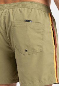 Khaki színű fürdőnadrág piros, sárga és barna függőleges csíkokkal. Hátsó zsebének patent és szemnek megfelelő részlete van. Könnyű anyagból készült.