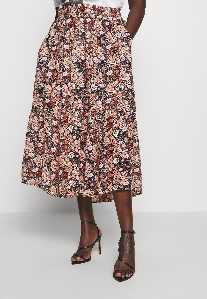 Personne portant un haut blanc, une jupe midi à motifs fleuris avec des poches et des sandales à talons noires à brides, debout devant un fond uni.