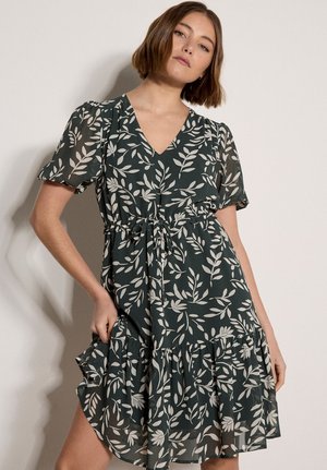 Frau trägt ein dunkelgrünes Kleid mit weißen Blattmustern, kurzen Puffärmeln, V-Ausschnitt und gerafftem Saum, posiert vor einem schlichten hellen Hintergrund.