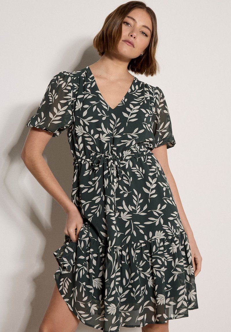 Femme portant une robe vert foncé avec des motifs de feuilles blanches, des manches courtes bouffantes, un décolleté en V et un ourlet volanté, posant devant un fond clair uni.