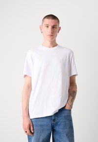 T-shirt en coton blanc avec un col rond et des manches courtes, arborant un logo discret sur la poitrine. Porté avec un jean en denim bleu.