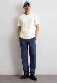 Witte t-shirt met korte mouwen en een klein logo op de borst, gecombineerd met donkerblauwe spijkerbroek en zwarte veterschoenen.
