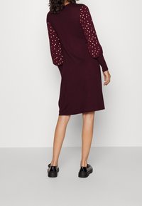 Robe bordeaux avec de longues manches à motifs comportant des points dorés. Texture lisse, design jusqu'au genou, associée à des chaussures à plateforme noires.