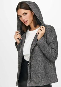 Giacca grigia con cappuccio in un tessuto texturizzato, chiusura con zip e due tasche laterali, indossata sopra una camicia bianca e pantaloni neri.