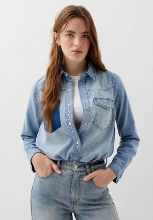 Světle modrá džínová košile s button-down přední částí, dekorativními nýty, dvěma náprsními kapsami a kontrastním tmavě modrým panelem na levé straně.