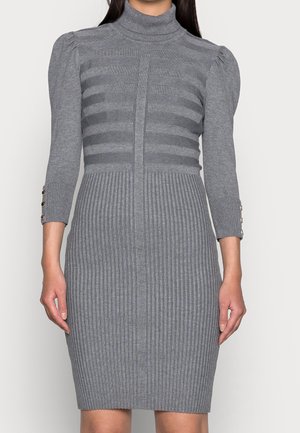 Frau trägt ein eng anliegendes graues, geripptes Strickkleid mit Puffärmel, dreiviertel Ärmeln und Knopfdetails an den Manschetten.