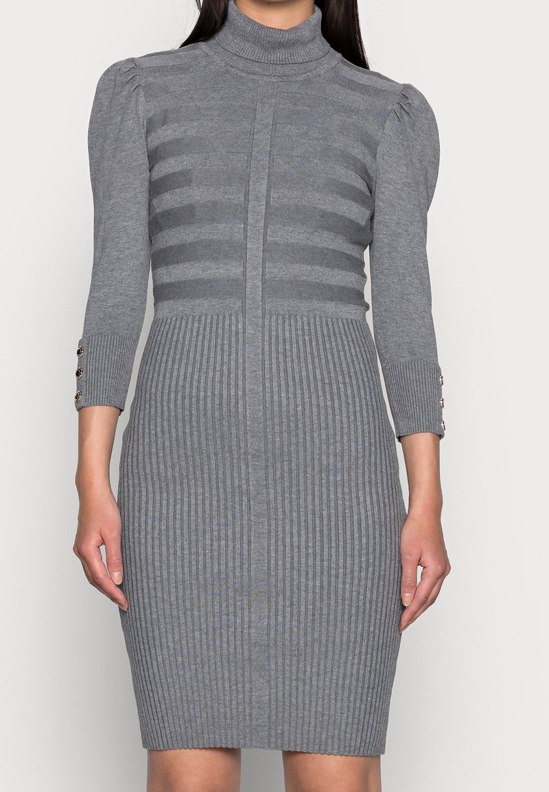 Femme portant une robe en maille côtelée grise cintrée avec des épaules bouffantes, des manches trois-quarts et des détails de boutons aux poignets.