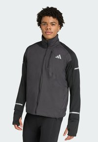 Black jacket featuring a zip front, high collar, reflective details, and matte finish. Long sleeves with thumbholes, made from lightweight fabric.