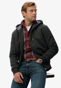 Chaqueta con capucha y cremallera en gris carbón, con capucha forrada de felpa, combinada con una camisa de cuadros roja, vaqueros azules y un cinturón marrón.