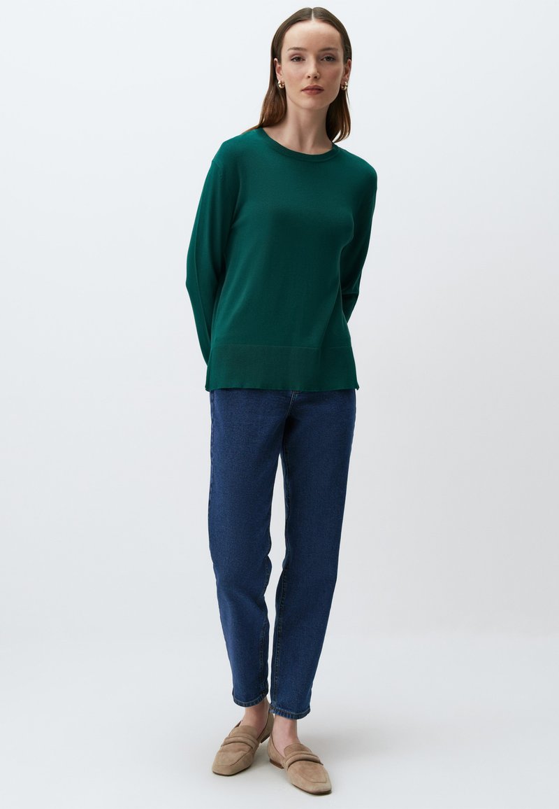 Zielony sweter dziany z okrągłym dekoltem, długimi rękawami i prostym dołem, w połączeniu z niebieskimi dżinsami i beżowymi mokasynami.