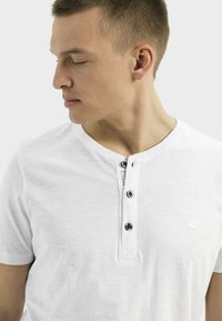 Witte short-sleeve shirt met een ronde hals, voorzien van een knoopsluiting met zwarte knopen en een klein logo op de borst. Zachte textuur.