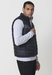 Schwarze Pufferweste mit einem Reißverschluss an der Vorderseite und hohem Kragen, kombiniert mit einem hellgrauen, strukturierten Langarmshirt und dunklen Jeans.