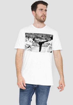 White cotton t-shirt featuring a black-and-white image of a martial arts kick. Short sleeves, crew neck, with a relaxed fit.