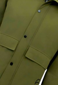 Olijfgroene jas met een gladde textuur, voorzien van zwarte drukknopen, twee borstzakken en een slanke silhouet.