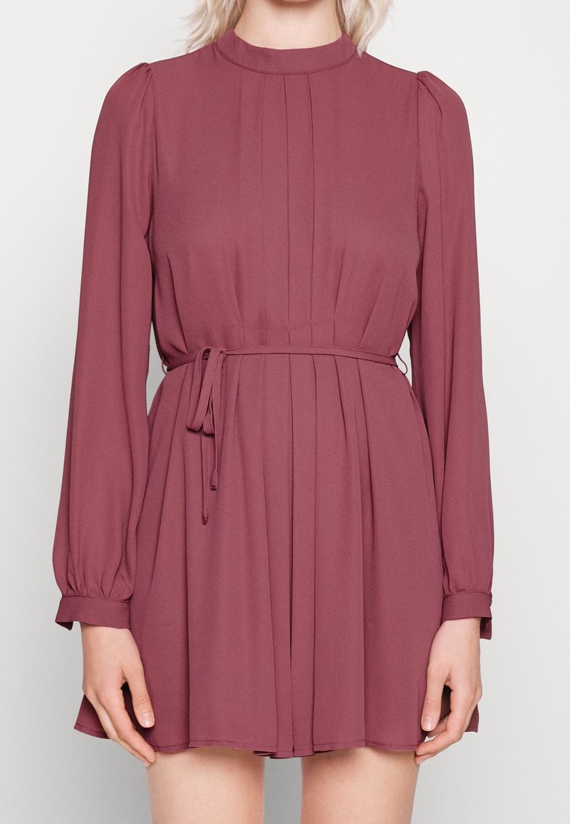 Femme portant une robe mauve à manches longues et col haut, avec une ceinture à la taille et une jupe plissée, debout devant un fond clair uni.