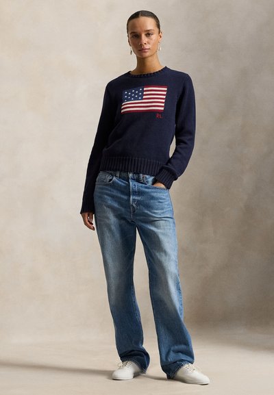 Navy knitted sweater with a red and white American flag design, paired with light blue straight-leg jeans and white sneakers.