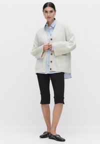 Beige oversized kofta med ribbad textur, knapplist fram, bärs över en ljusblå randig skjorta, tillsammans med svarta knä-långa shorts och svarta platta skor.
