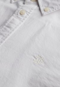 Camisa blanca con cuello, botones beige y un pequeño logo blanco bordado cerca de la zona del pecho.