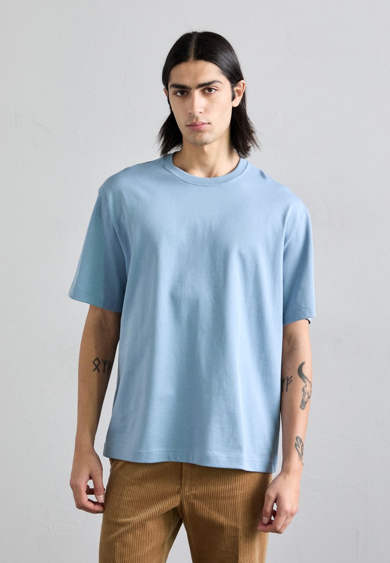 Camiseta de manga corta azul claro, hecha de algodón, con un escote redondo y un corte holgado, combinada con pantalones de pana marrones.