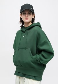 Person med mørkegrønn Nike hettegenser og svart Nike caps, står med hendene i hettegenserlommen og ser litt til siden.