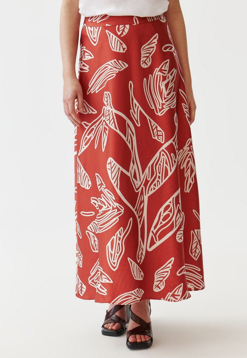 Jupe maxi en tissu rouge avec des motifs de feuilles abstraites blancs. Équipée d'une taille cintrée, elle tombe jusqu'à la cheville. Portée avec des sandales noires à lanières.